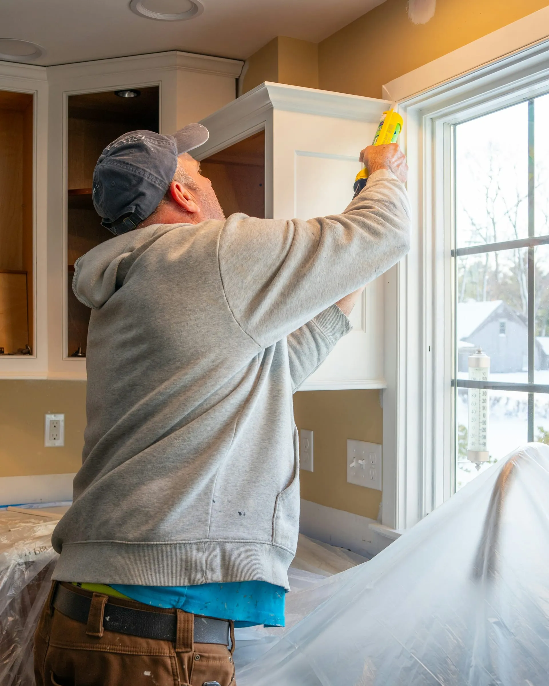 A handyman sealing a kitchen cabinet by a bright window