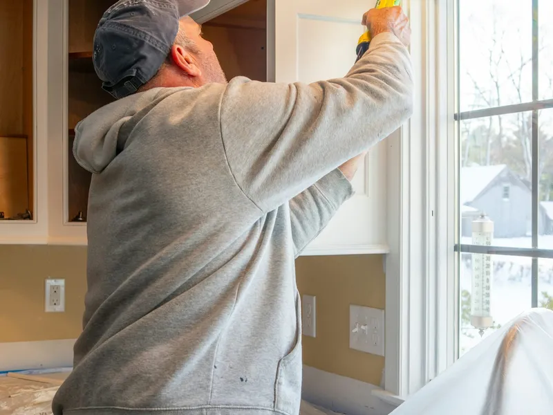 A technician sealing a kitchen cabinet by the window