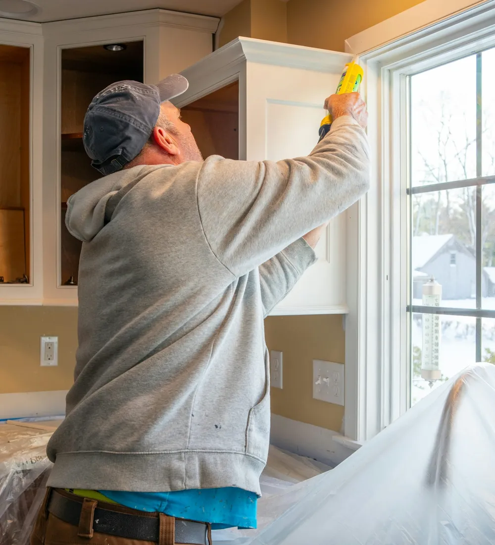 A handyman sealing a kitchen cabinet by a bright window